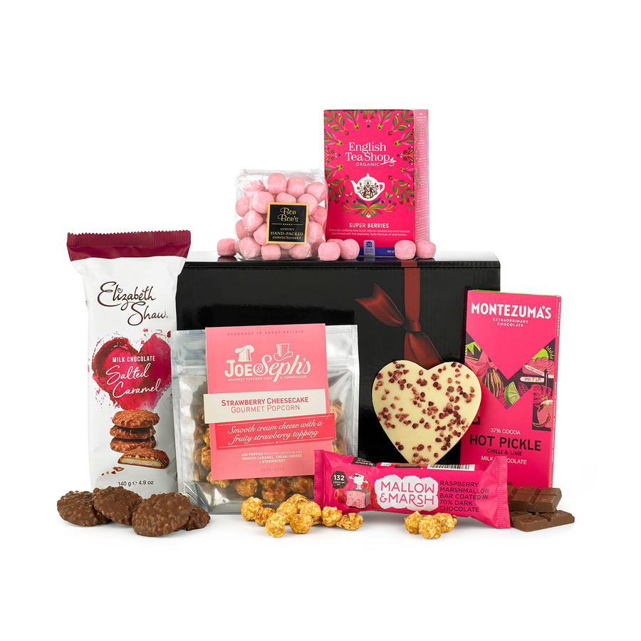 The Made With Love alcohol free gift hamper laid out on a white table top. Including Marshmallow, Hot Pickle Chilli & Lime chocolate bar, Salted caramel milk chocolate biscuits, strawberry popcorn & more all neatly hand-packed int0 the black gift box pictured with a ruby red ribbon tied around it.