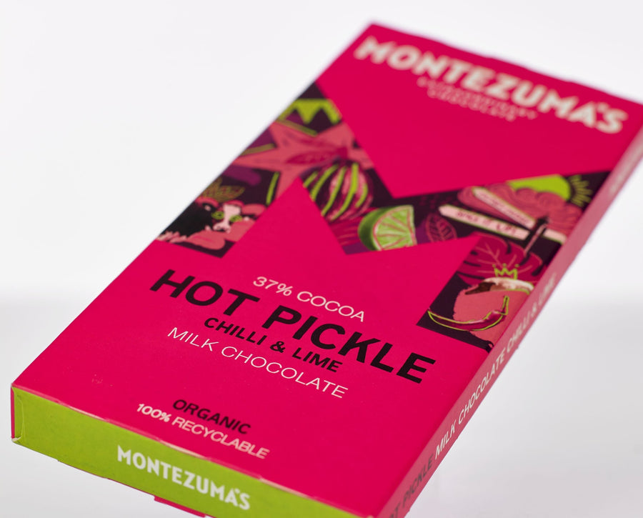 Montezuma hot pickle chilli & lime milk chocolate bar pictured on a white table top. The bright pink and green packaging are featured heavily in the image. 